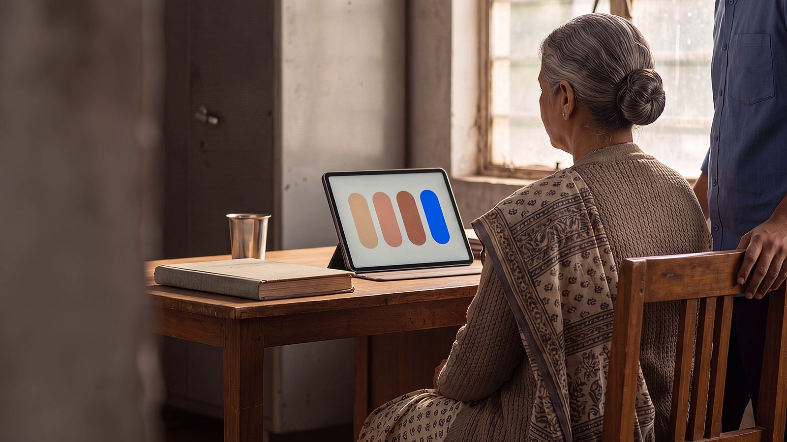 A grey-haired woman in a saree sits at a wooden desk in front of a tablet showing four soft coloured bars, a closed cloth-bound register and a steel tumbler beside her, while a colleague rests a hand on her chair.